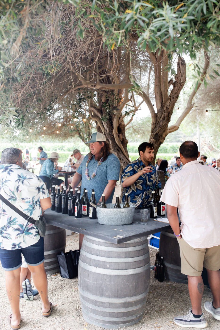 A lush outdoor wine tasting event under a large tree with people sampling wines at Kaena Wine Co. during a wine club gatherin.