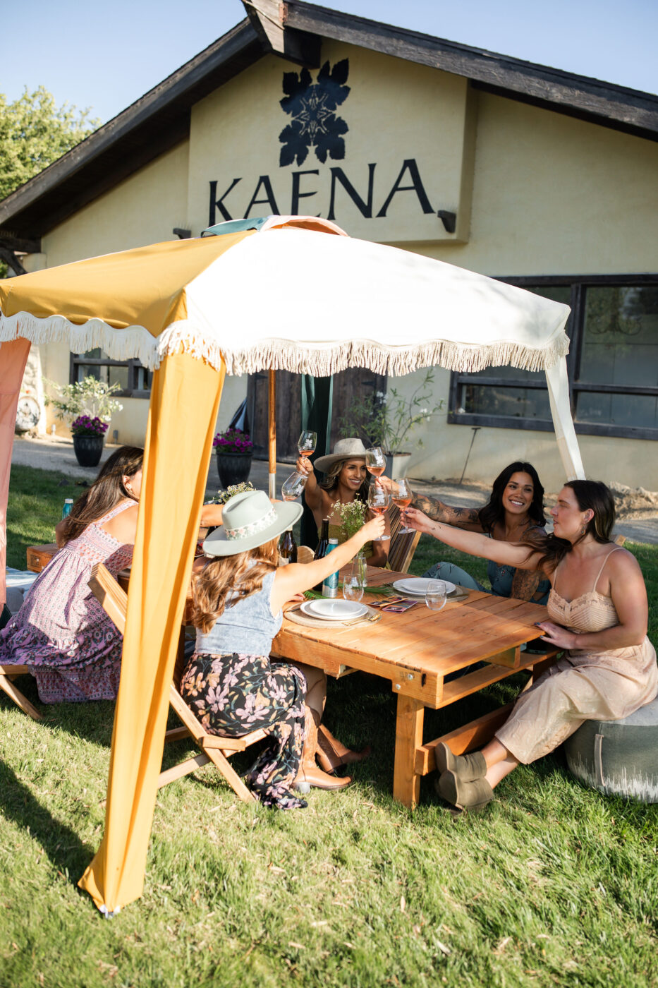 Elegant group of women enjoying a private outdoor event at Kaena Wine Co, seated around a wooden table under a yellow tent.