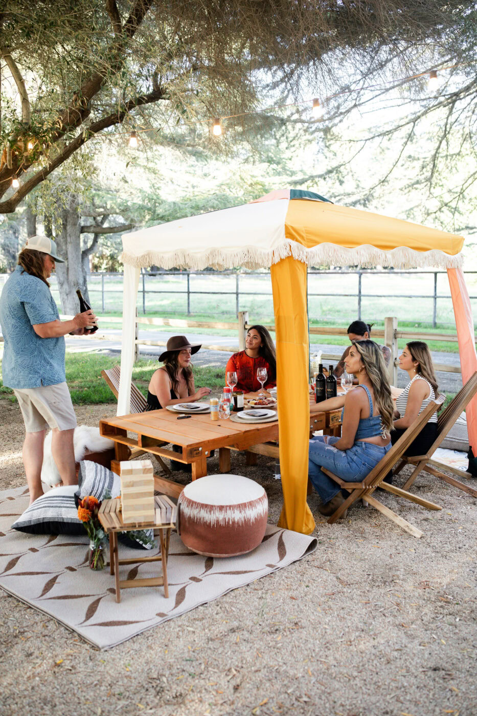 A lively outdoor gathering of friends enjoying wine under a yellow and white canopy in a lush, shaded park setting.
