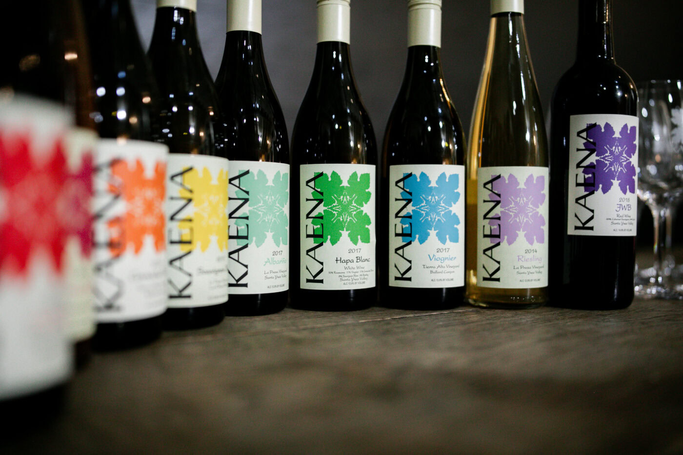 Kaena Wine Co bottles with colorful labels on a wooden surface, showcasing a variety of wines from the Kaena Wine Company.