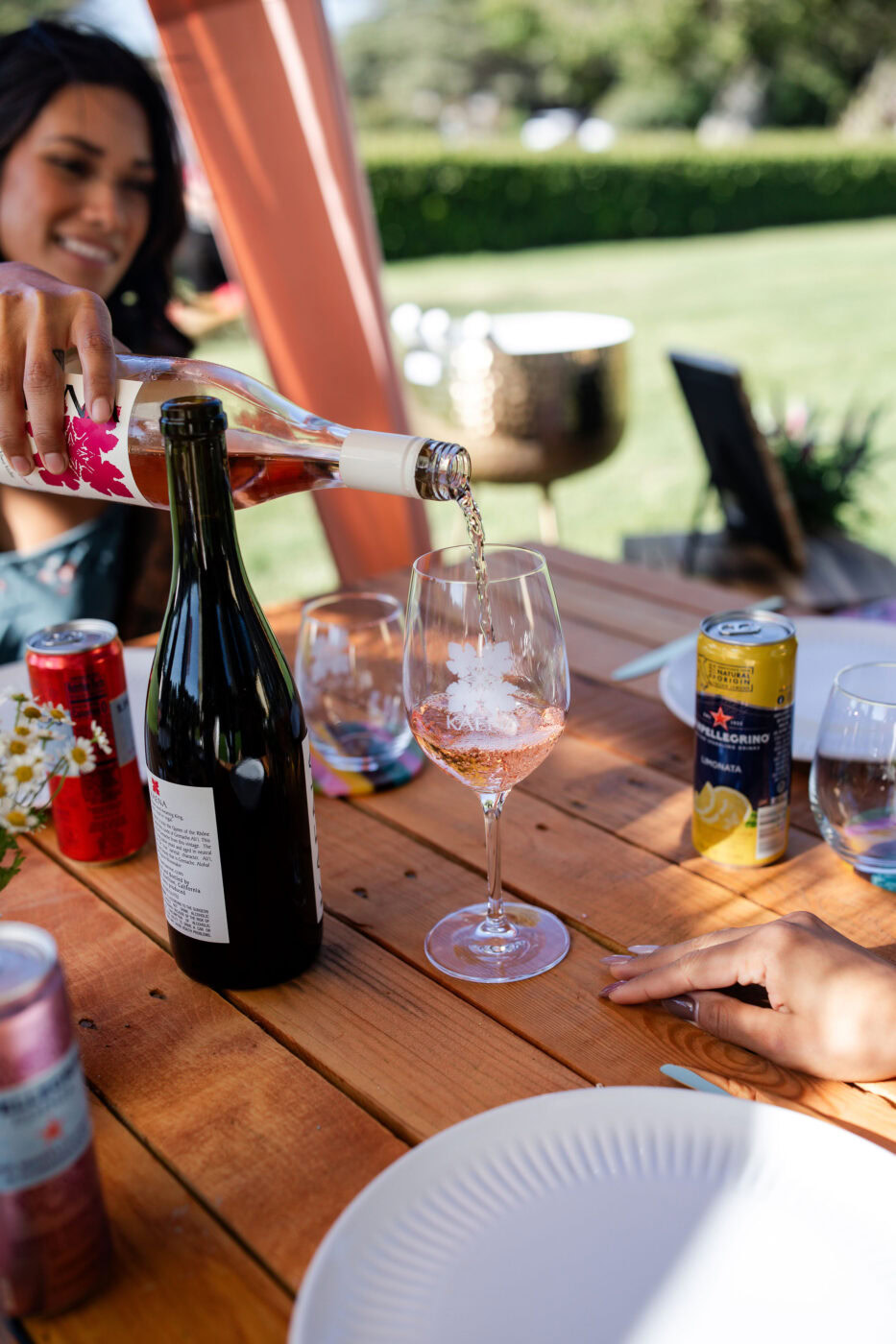 A woman pouring rosé wine into a glass at an outdoor gathering with wine bottles and cans on a wooden table.