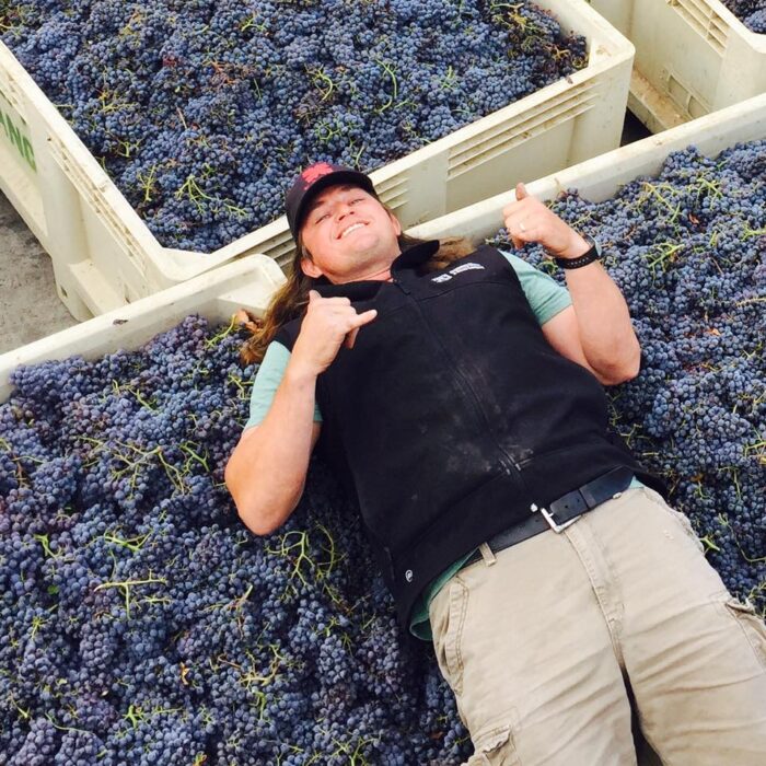 Kaena winemaker lying on purple grapes in a vineyard, smiling and making a thumbs-up gesture. Best Wines In Santa Barbara County