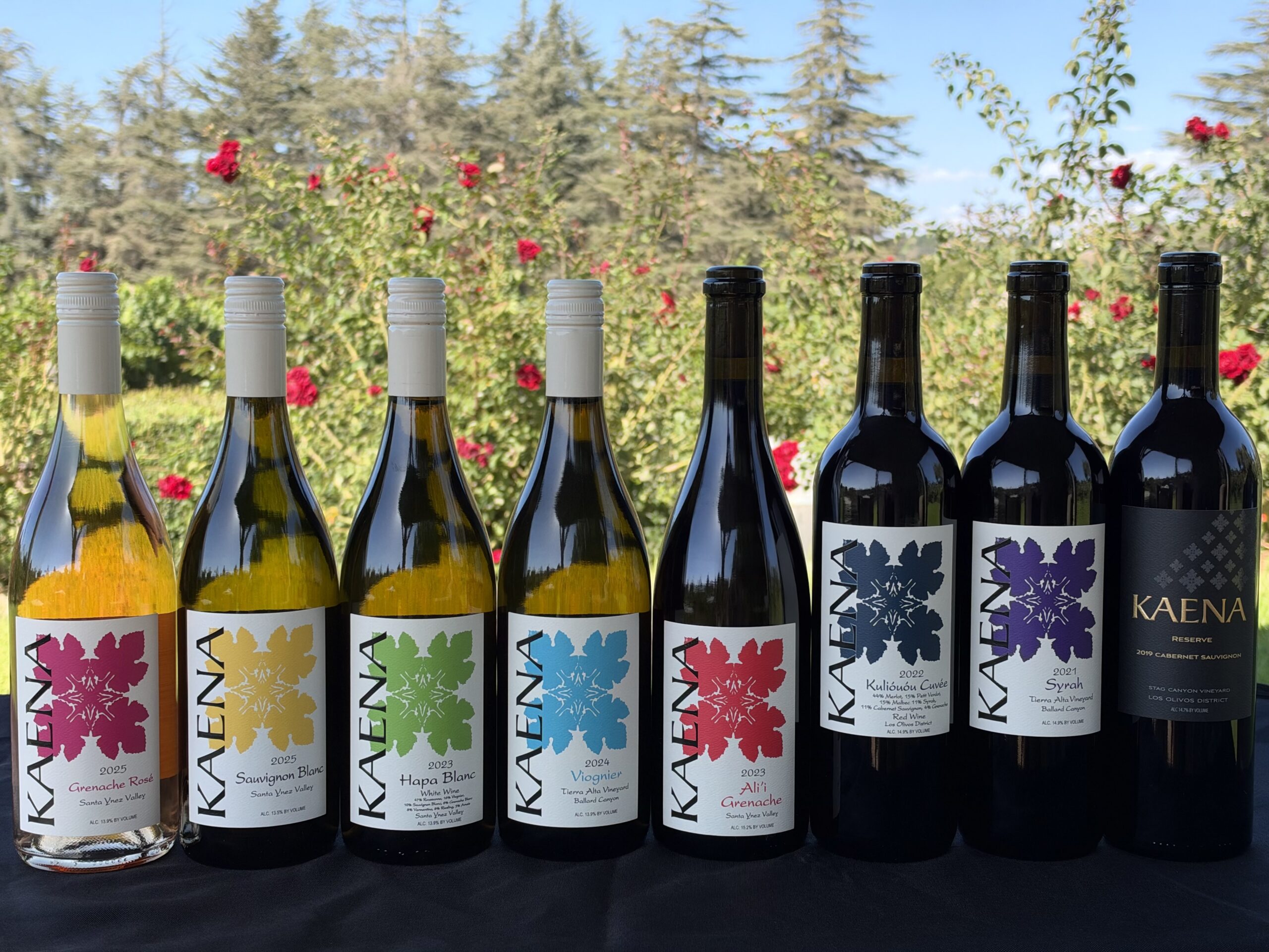 Kaena Wine Co bottles displayed outdoors with a garden background of roses and trees.